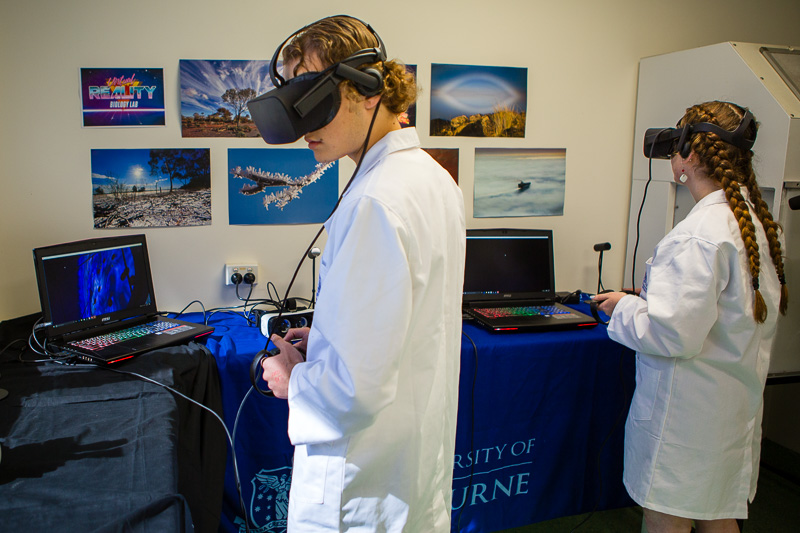 Two students in white lab coats wear virtual reality headsets to explore their work