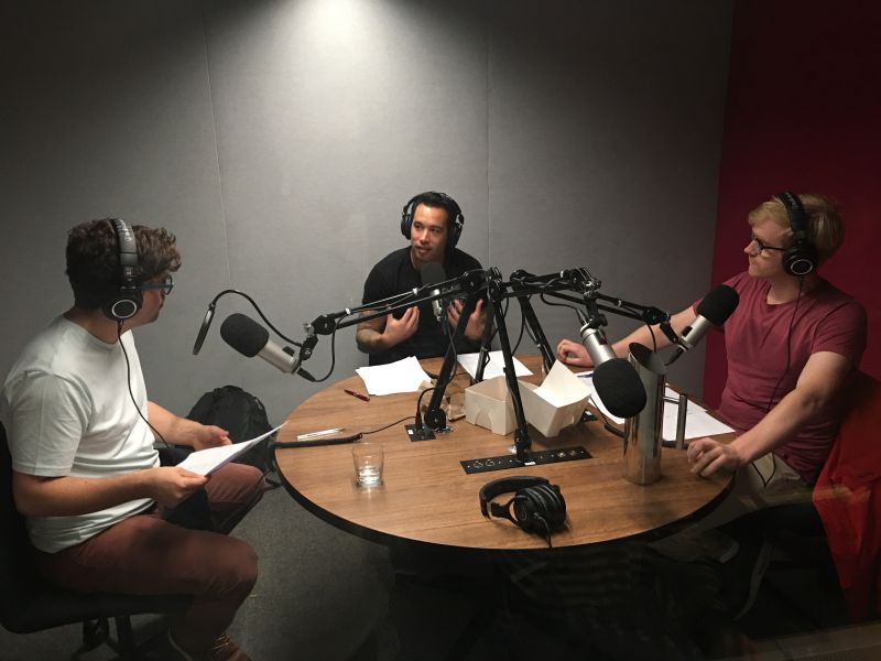 Three men sit around a table in a podcast studio wearing headphones and having an animated discussion.