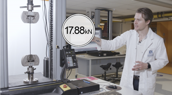 Presenter in a lab coat stands beside machinery, gesturing to an overlaid graphic measurement that hovers above.