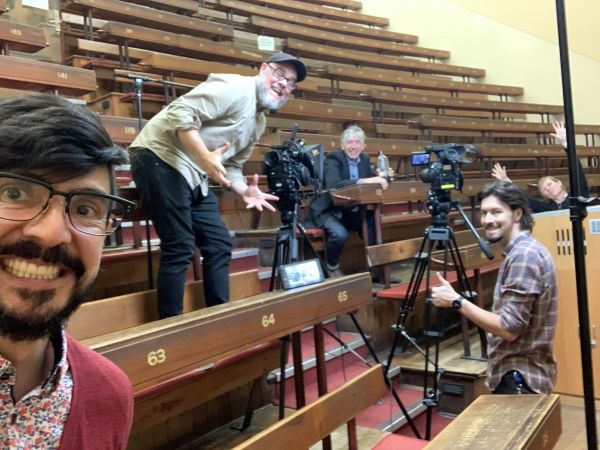 Behind the scenes in an old lecture theatre, the producer takes a selfie with another crew member and the presenter.