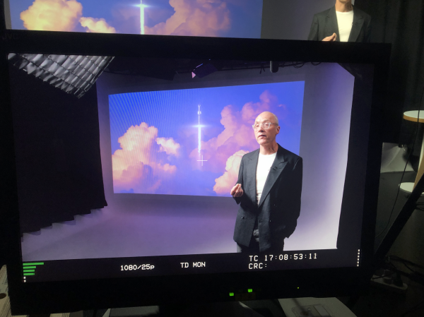 A presenter standing in a studio in front of a screen with clouds can be seen through the viewing screen of a camera